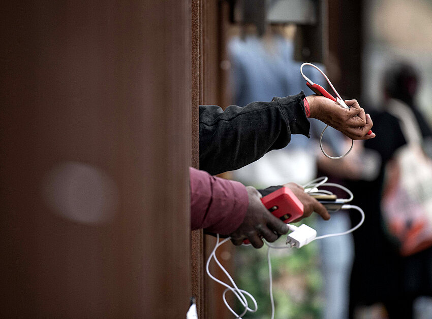 Solicitantes de asilo sostienen sus teléfonos a través del muro fronterizo mientras suplican a la gente que elijan su teléfono para ser cargado